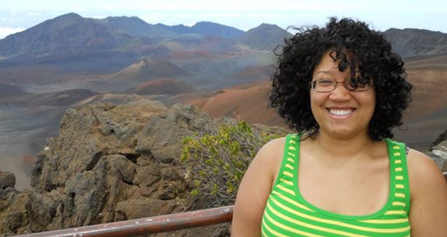 A woman smiles in front of a mountainous landscape with scattered vegetation.